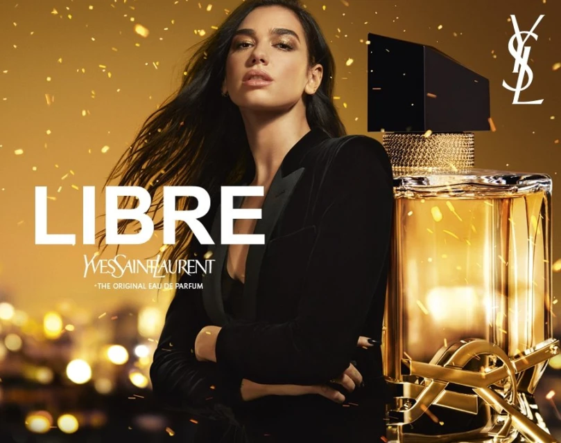 Dua Lipa poses in a black suit jacket next to a YSL Libre perfume bottle against a gold bokeh background in a beauty advertising shot.
