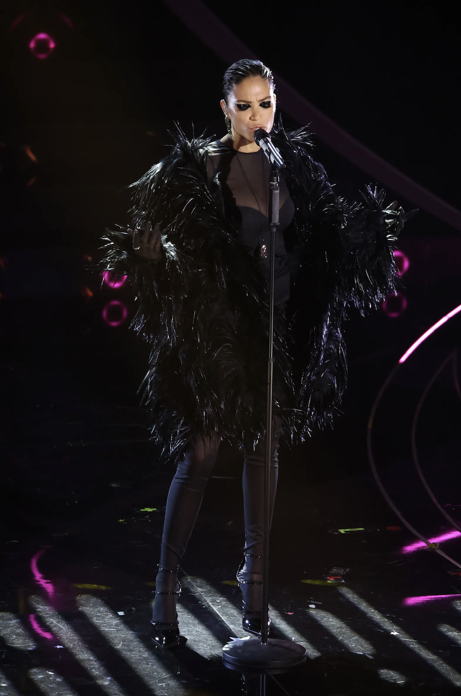 Adult woman in a feathered coat singing on stage into a microphone, with dynamic lighting and a concert atmosphere.
