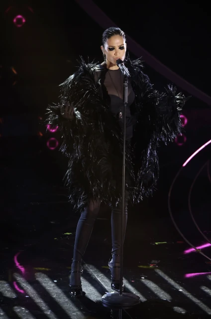 Adult woman in a feathered coat singing on stage into a microphone, with dynamic lighting and a concert atmosphere.