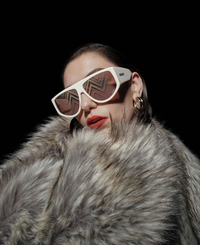 Close-up portrait of a woman wearing thick white sunglasses and a voluminous grey faux fur coat against a black background.