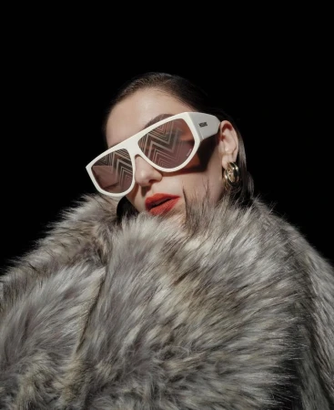 Close-up portrait of a woman wearing thick white sunglasses and a voluminous grey faux fur coat against a black background.