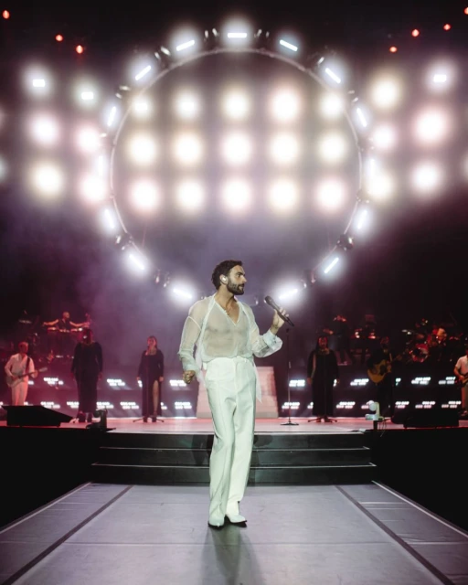 Male musician performs on stage with band and bright lighting. He wears a white outfit, holding a microphone, with stage lighting.