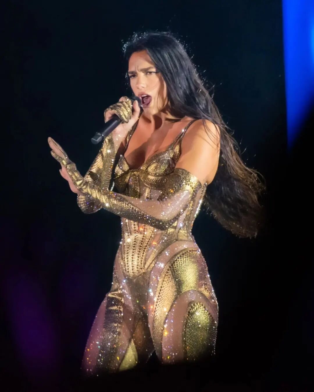Dua Lipa performing on stage with a microphone, wearing a gold sequined outfit, with a dark background and stage lighting.