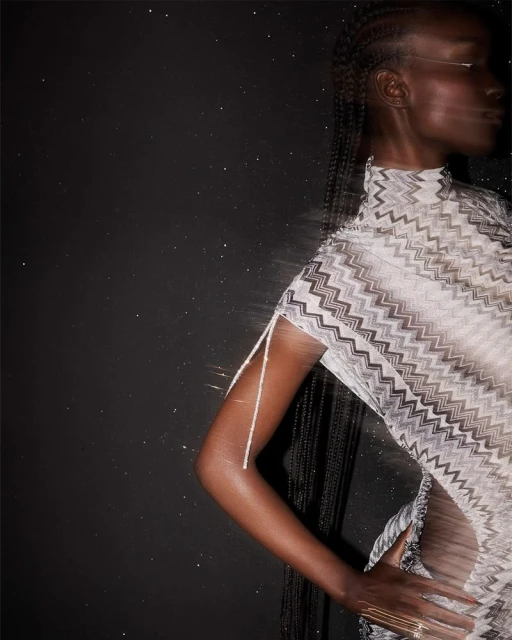 Woman with dark skin and braided hair poses in a zig-zag pattern dress against a dark background, showcasing fashion and style.