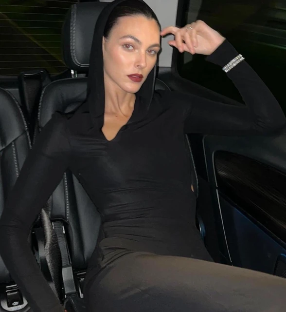 Adult woman with dark hair wearing a long sleeve black hooded top poses in a car interior with black leather seats.