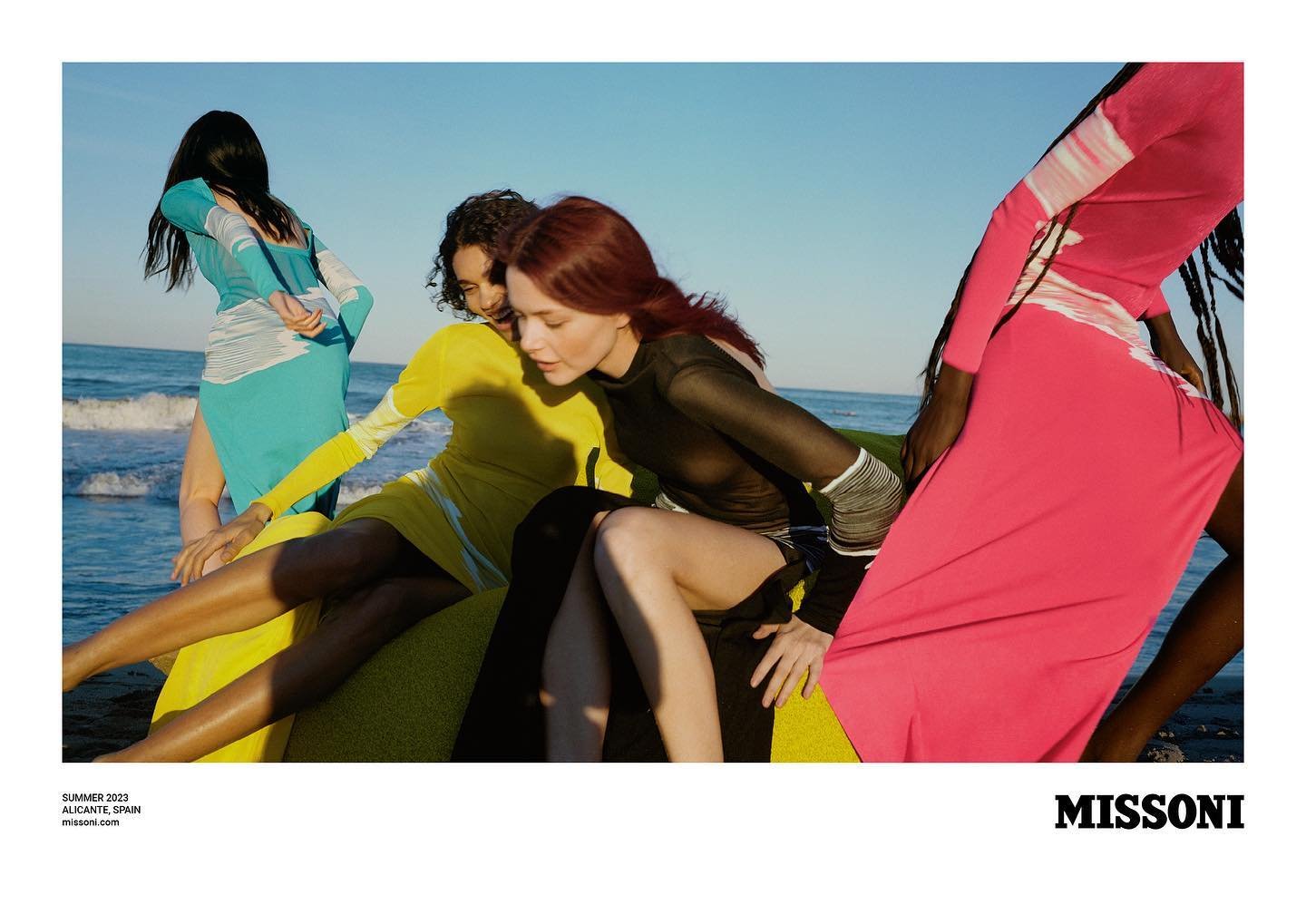 Fashion editorial shot of four adult female models in colorful long sleeve dresses posing on the beach with ocean and sky background.
