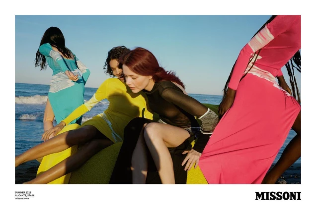 Fashion editorial shot of four adult female models in colorful long sleeve dresses posing on the beach with ocean and sky background.