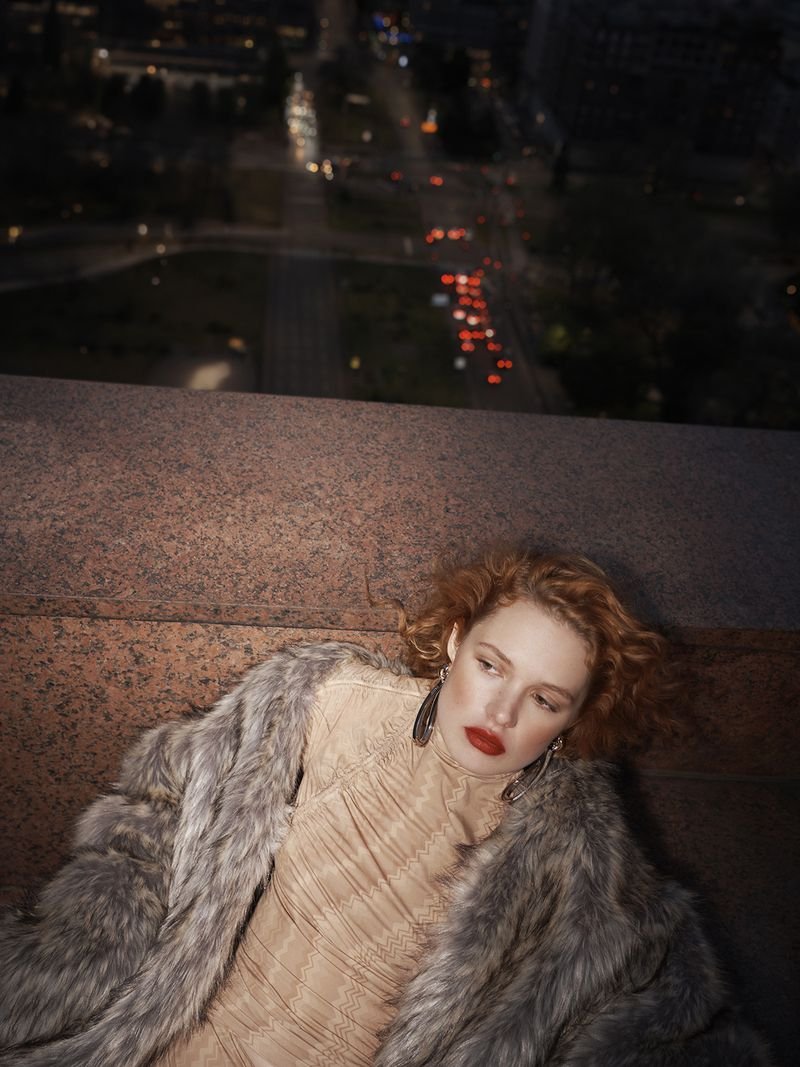 Woman with red lipstick and curly red hair wearing a fur coat and dress lying on a stone surface with a city view in the background.