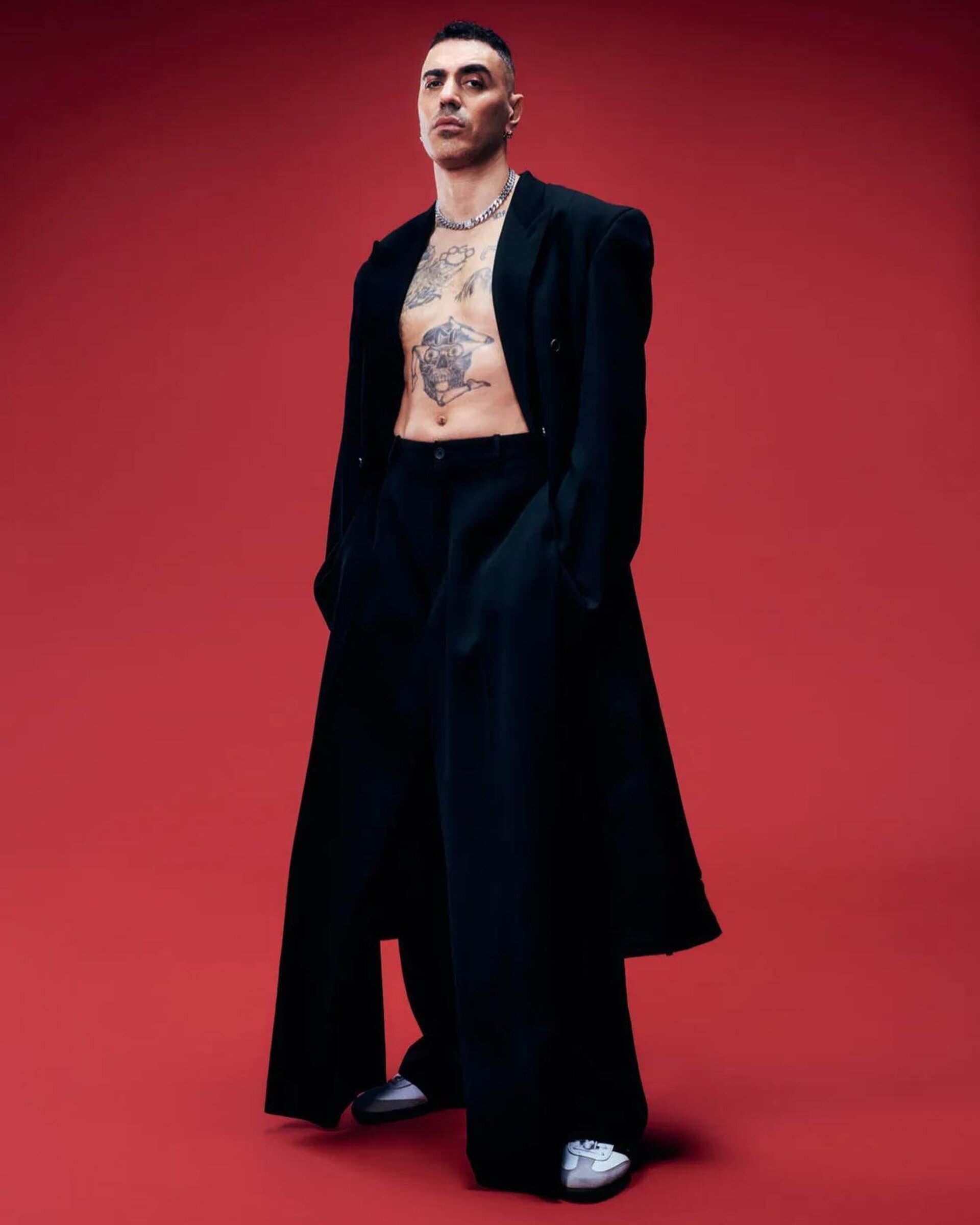 Stylish man in oversized black coat and pants poses confidently against a vibrant red backdrop, showcasing tattoos and silver chain.
