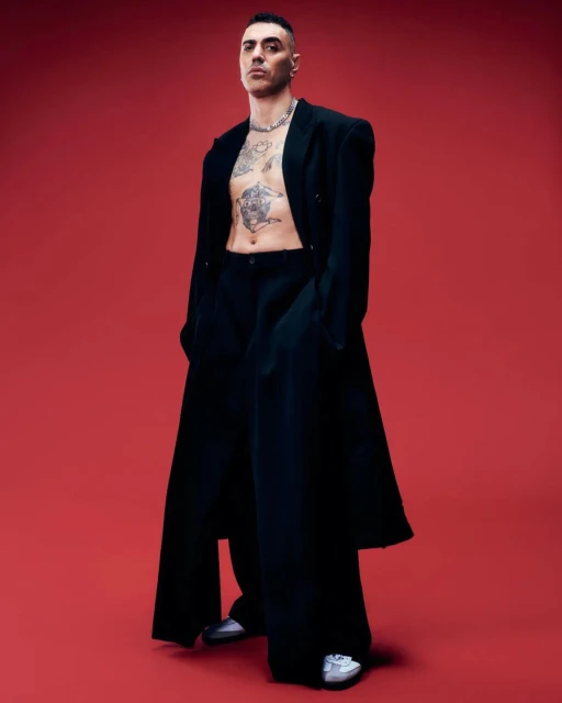 Stylish man in oversized black coat and pants poses confidently against a vibrant red backdrop, showcasing tattoos and silver chain.