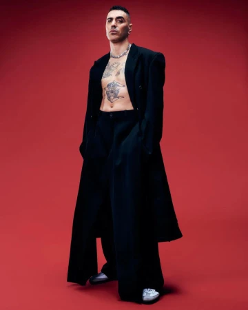 Stylish man in oversized black coat and pants poses confidently against a vibrant red backdrop, showcasing tattoos and silver chain.