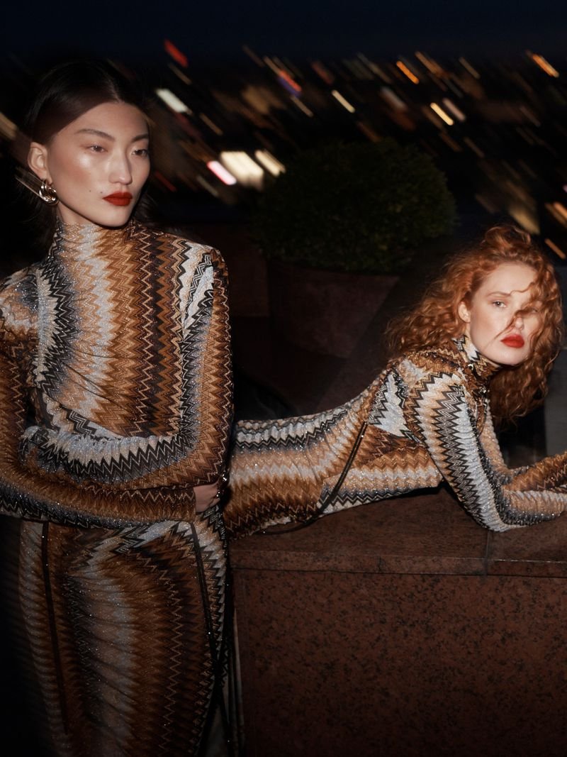 Two women in patterned dresses pose on a rooftop at night with city lights in the background, fashion and style photography.