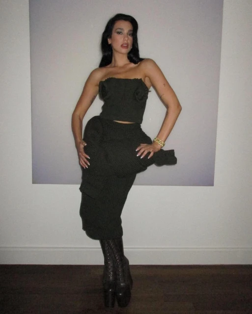 A woman in a dark green outfit posing in front of a white wall with a picture in the background in an indoor setting.