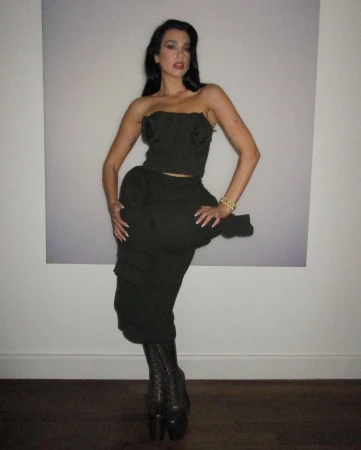 A woman in a dark green outfit posing in front of a white wall with a picture in the background in an indoor setting.