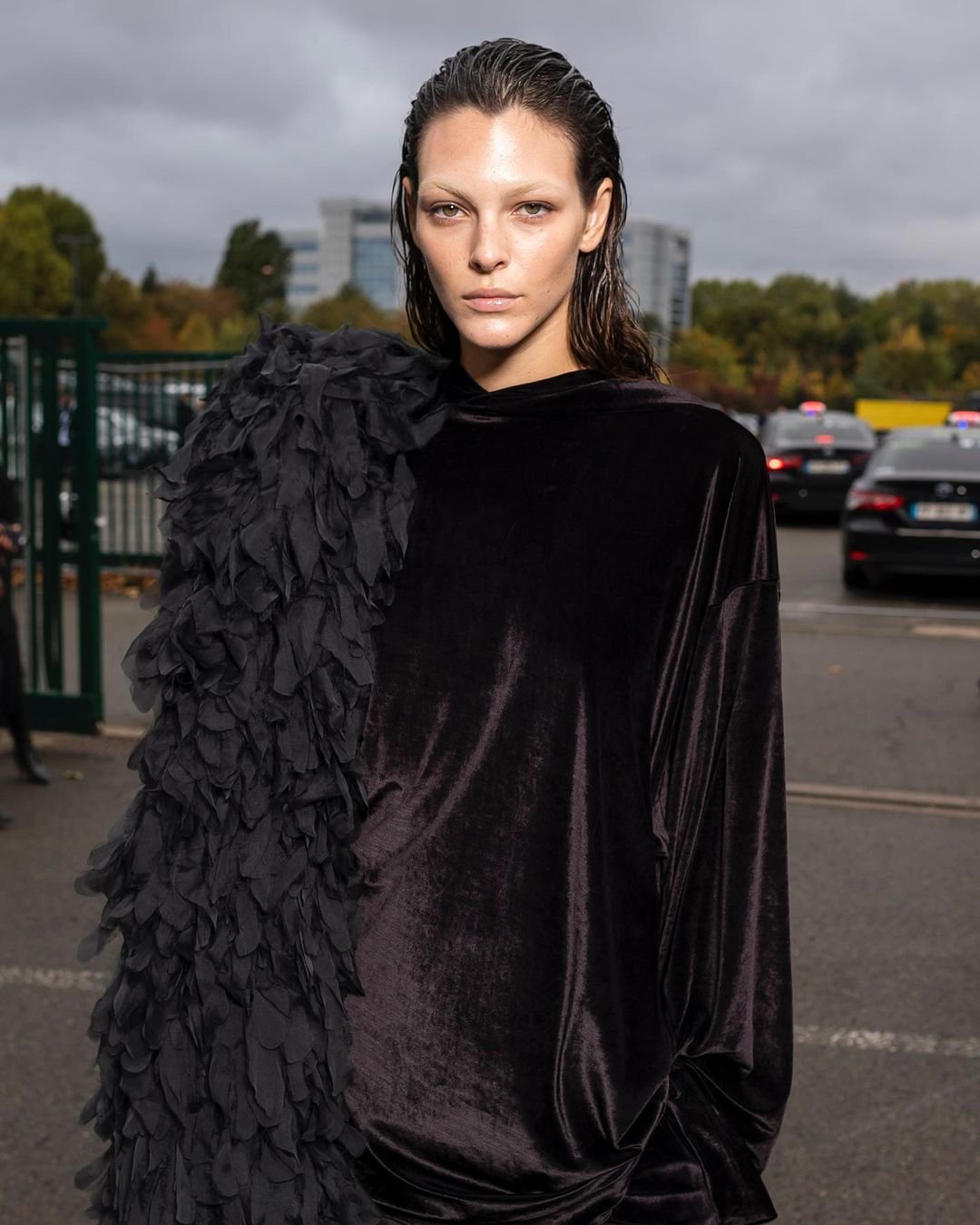Fashion model poses outdoors wearing black velvet hooded dress with feathered embellishments, with a blurred street and building background.