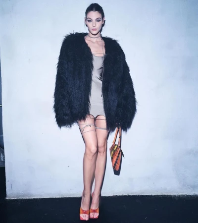 Model wearing a black faux fur coat and beige dress with a handbag against a plain grey wall