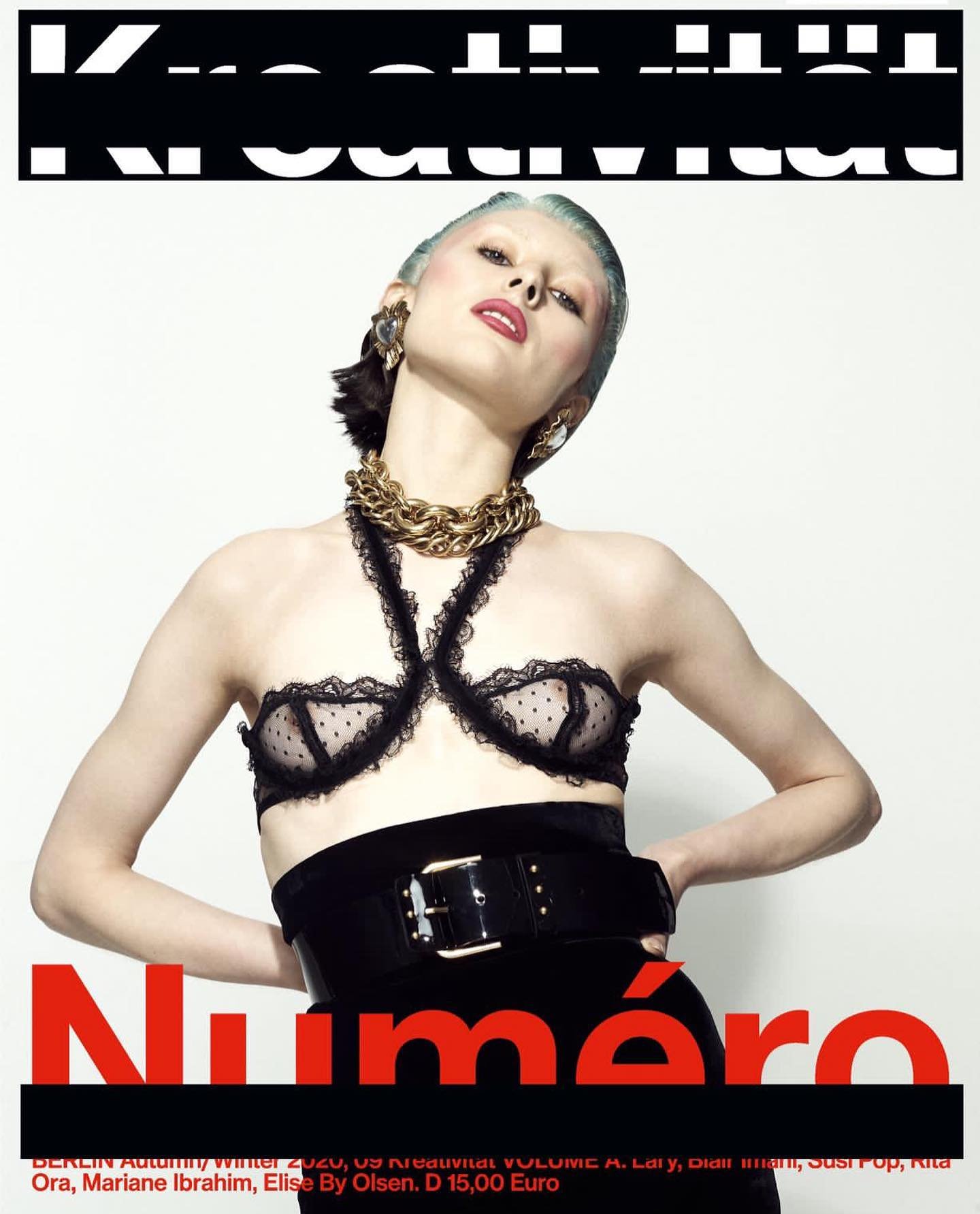 A woman with pale skin and blue hair poses on a magazine cover in a black bra and belt with a gold chain and a red title.