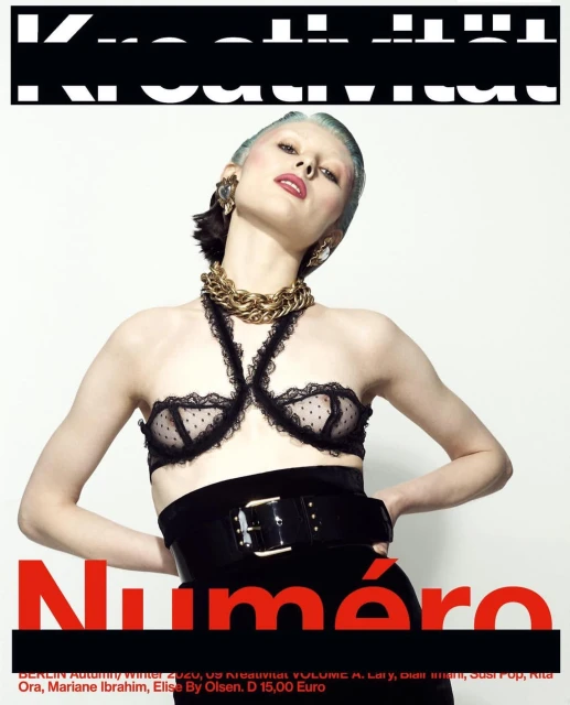 A woman with pale skin and blue hair poses on a magazine cover in a black bra and belt with a gold chain and a red title.