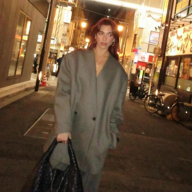 Adult female with auburn hair in oversized gray suit, carrying a woven bag, walking in a narrow urban street at night.