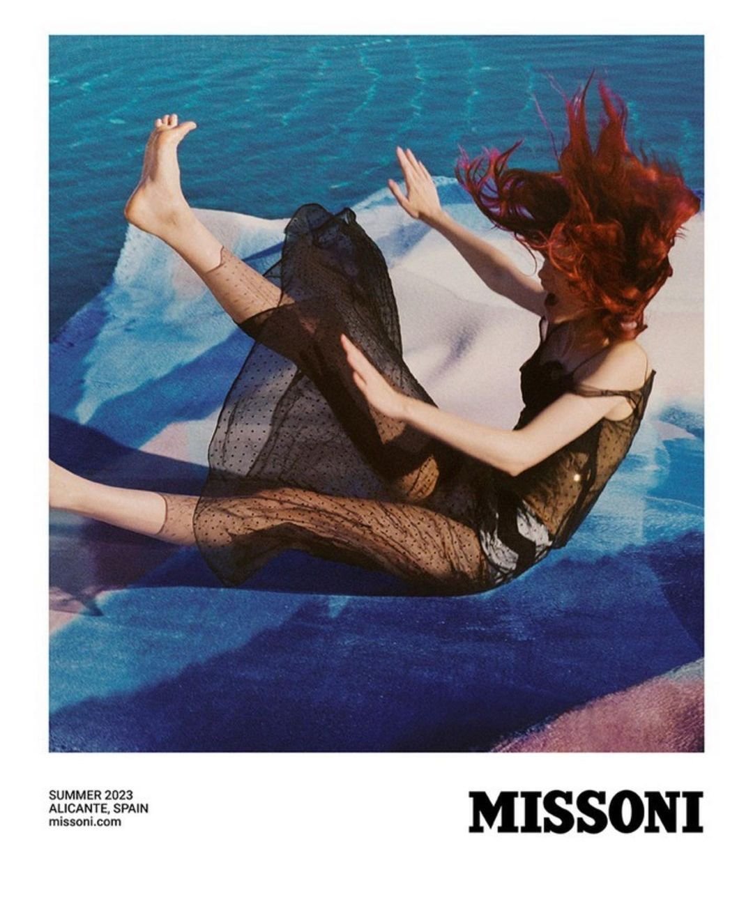 A woman with vibrant red hair poses dynamically in a sheer dress on a textured surface, set against a blurred blue and white backdrop.