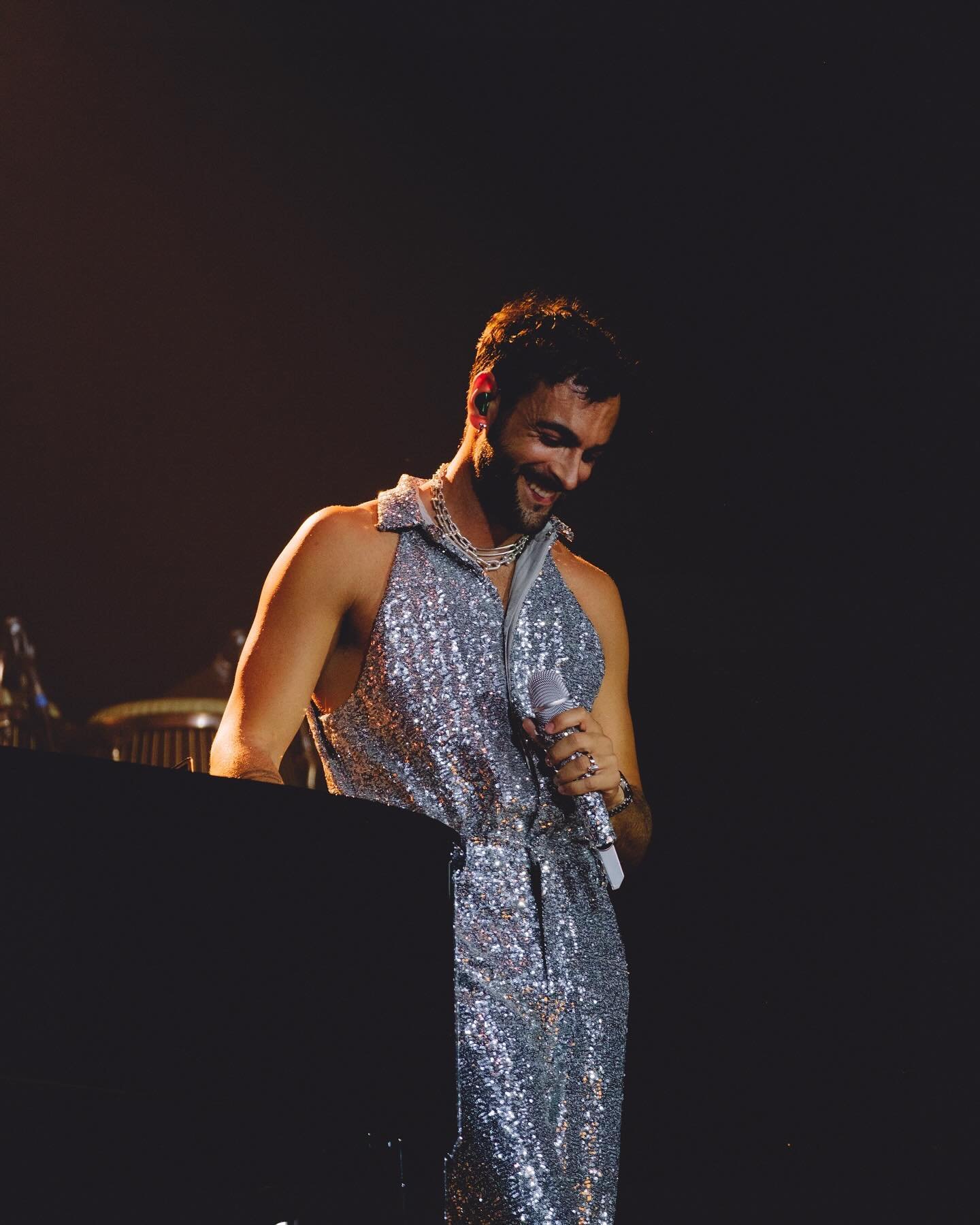 Man on stage singing, wearing a sparkly silver outfit and necklace, holding a microphone, with dark hair and a dark background.