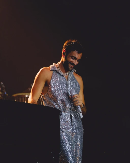 Man on stage singing, wearing a sparkly silver outfit and necklace, holding a microphone, with dark hair and a dark background.