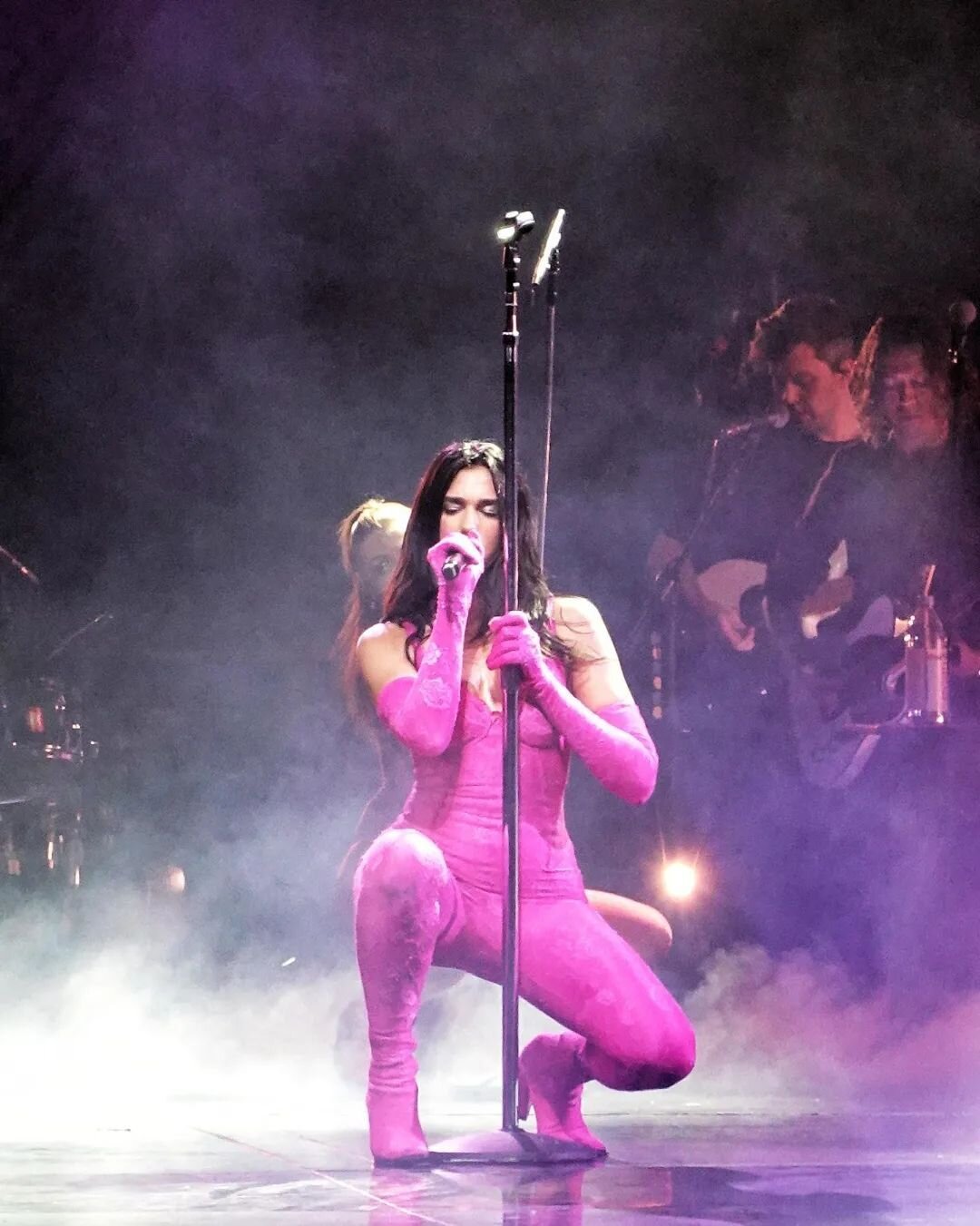 Dua Lipa performs live in pink bodysuit and platform boots, singing into microphone on stage with band in smoky background.