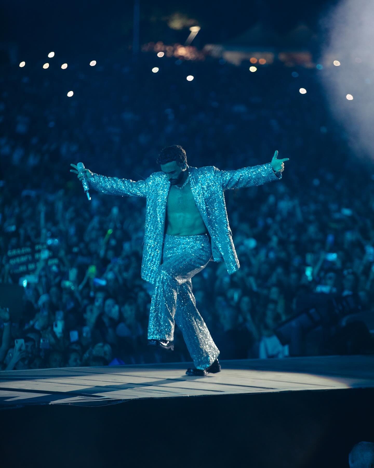 Performance shot of a male singer in a sparkly silver suit singing and gesturing on stage to a large crowd and bright lighting.