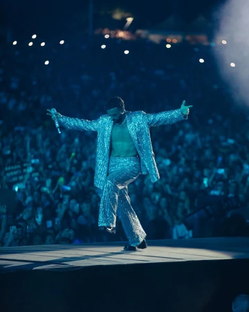 Performance shot of a male singer in a sparkly silver suit singing and gesturing on stage to a large crowd and bright lighting.