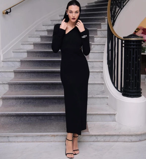 Elegant woman in a long black dress and strappy heels poses on marble stairs with an ornamental iron railing and floral decor in the background.