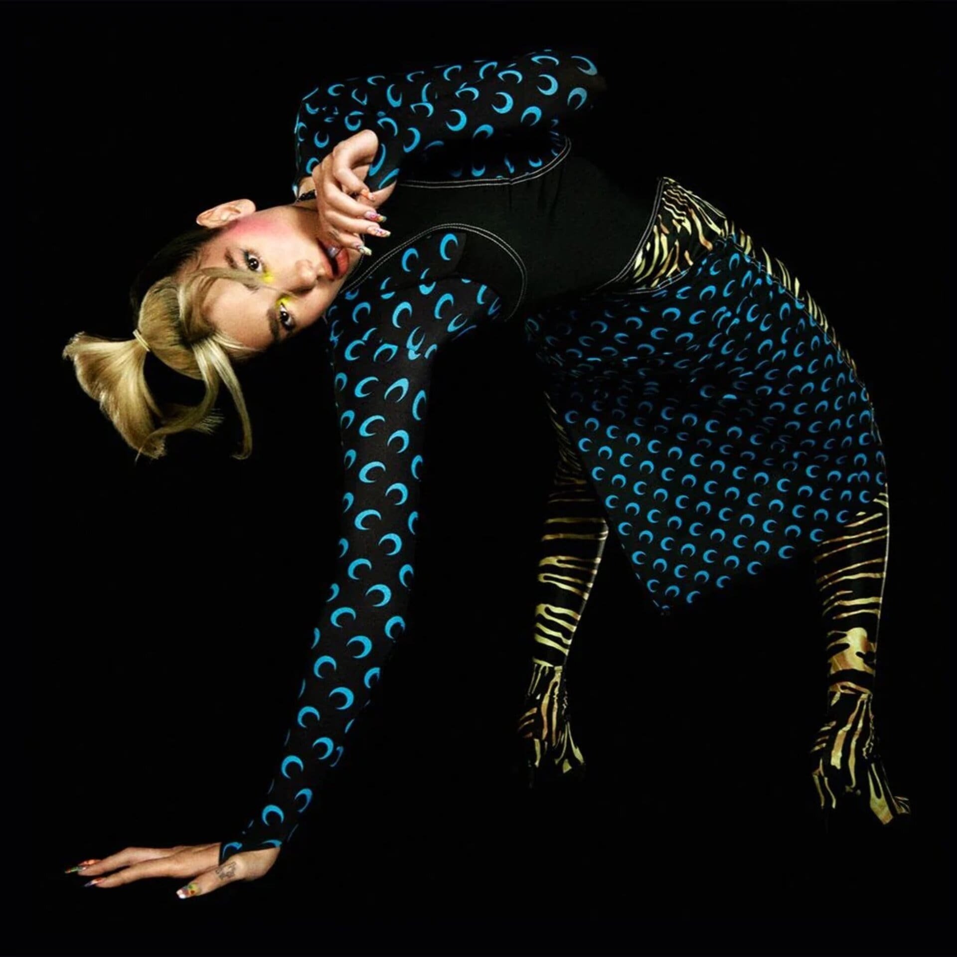 Woman in a crescent-moon print bodysuit and skirt bends over with one hand on the floor in a black studio backdrop.