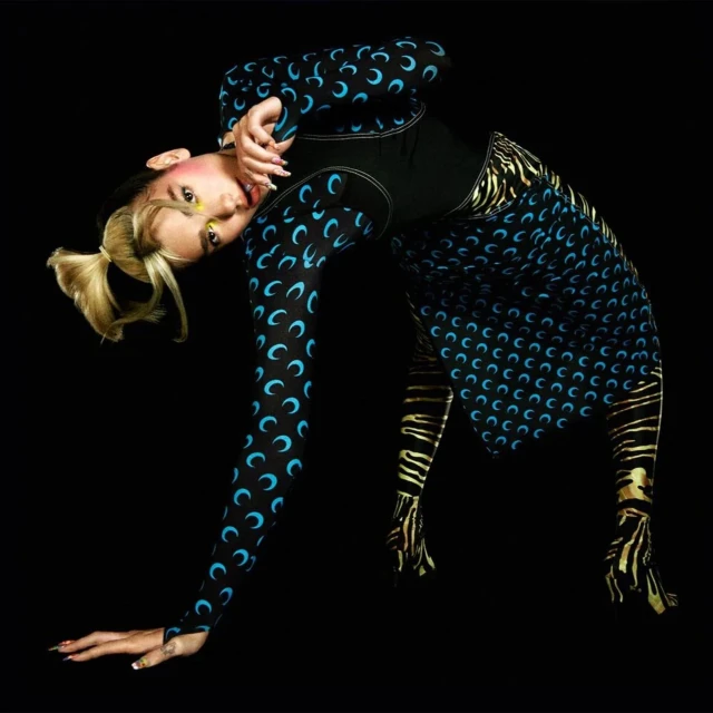 Woman in a crescent-moon print bodysuit and skirt bends over with one hand on the floor in a black studio backdrop.