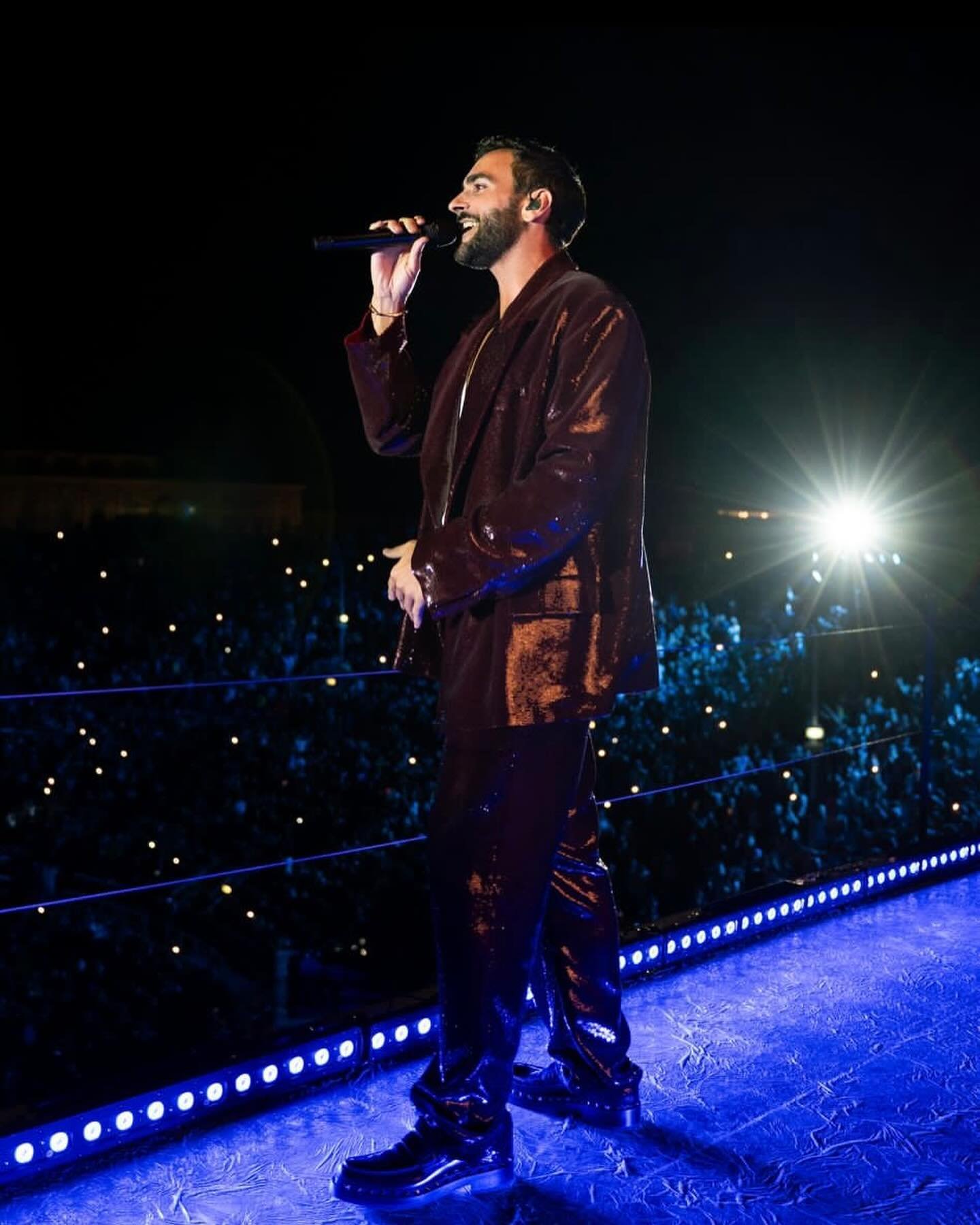 A bearded adult man sings on stage in a dark maroon, sequined suit with a microphone during a concert with bright stage lighting and a crowd.