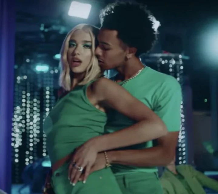 Two people in green outfits stand close together indoors, with a woman with blonde hair and a man with an afro.