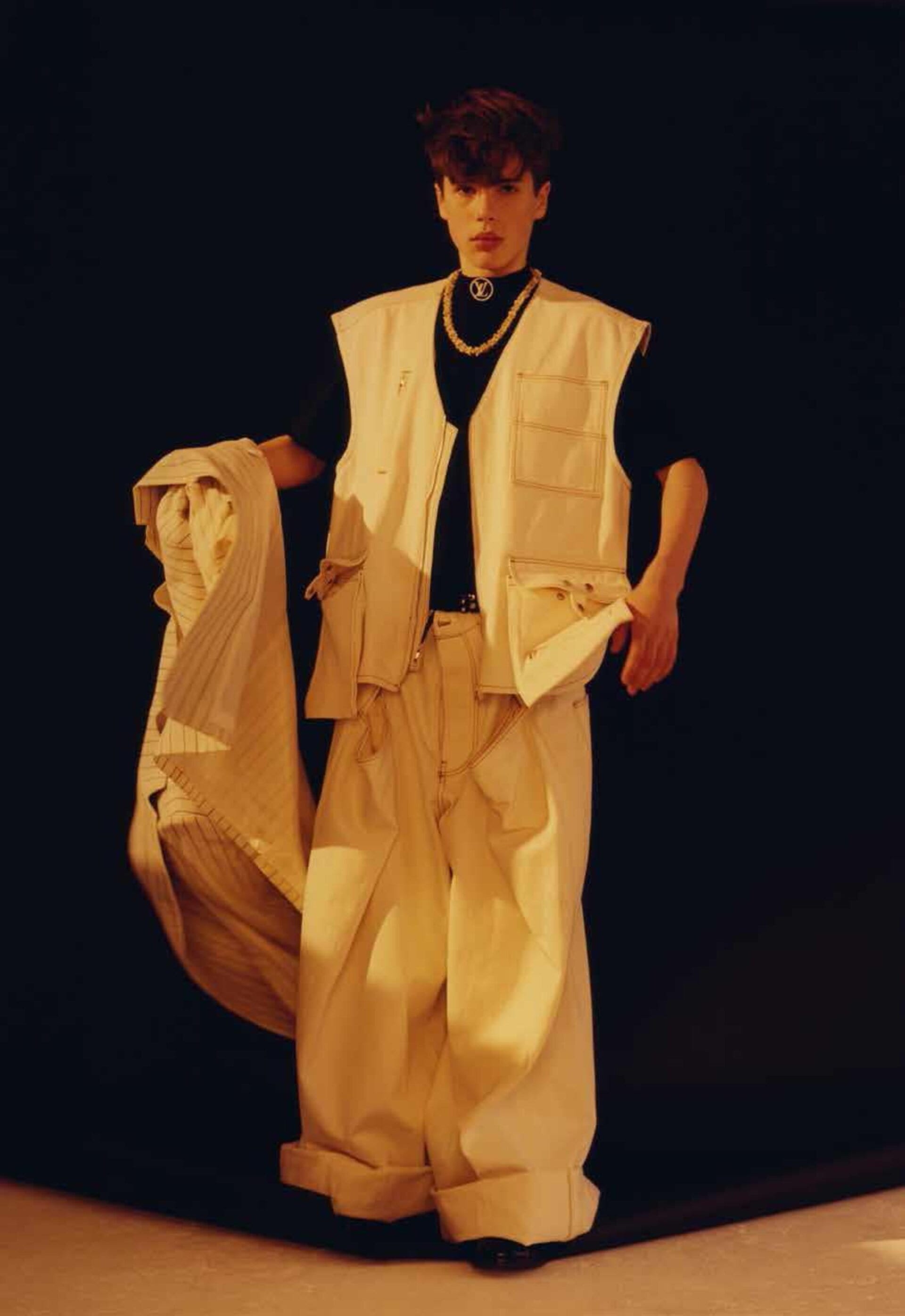 A young adult male model poses in a sleeveless beige outfit with loose pants against a dark backdrop, holding a striped jacket.