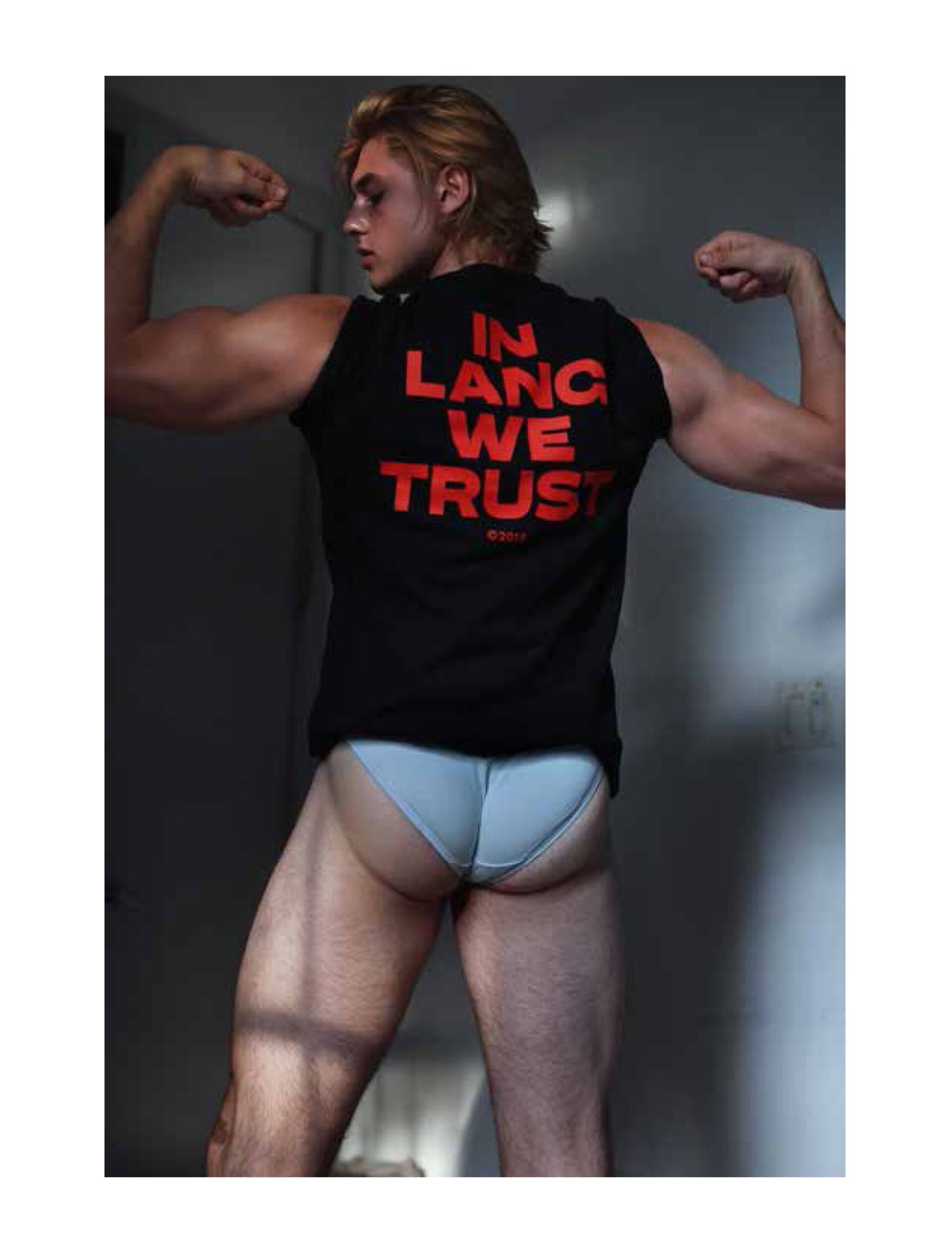 Blond male flexing arms in a sleeveless black shirt with red text and light blue underwear, indoors with light and shadow play.