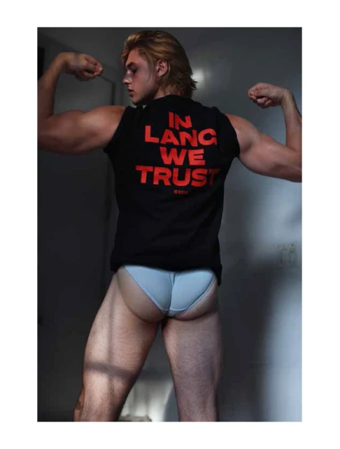 Blond male flexing arms in a sleeveless black shirt with red text and light blue underwear, indoors with light and shadow play.