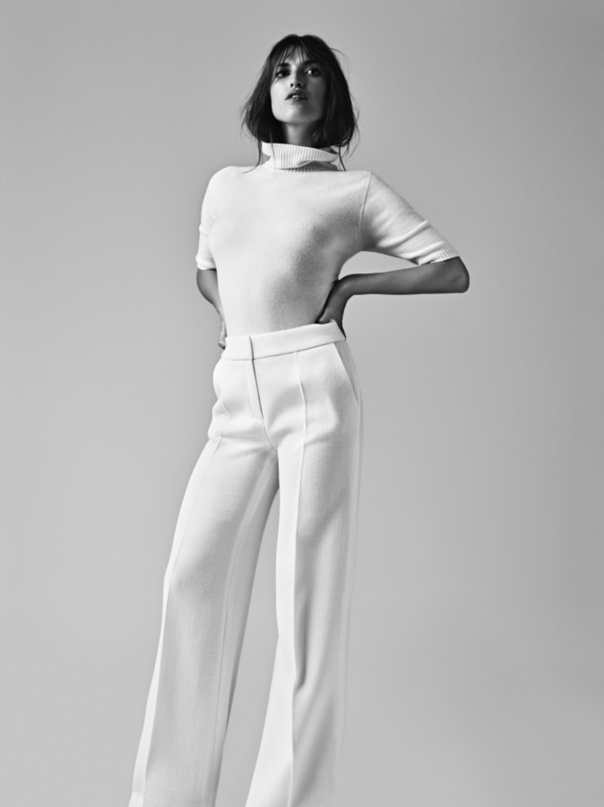 A slender adult woman poses in a photo studio, wearing a white turtleneck and matching high-waisted, wide-leg trousers in monochrome.