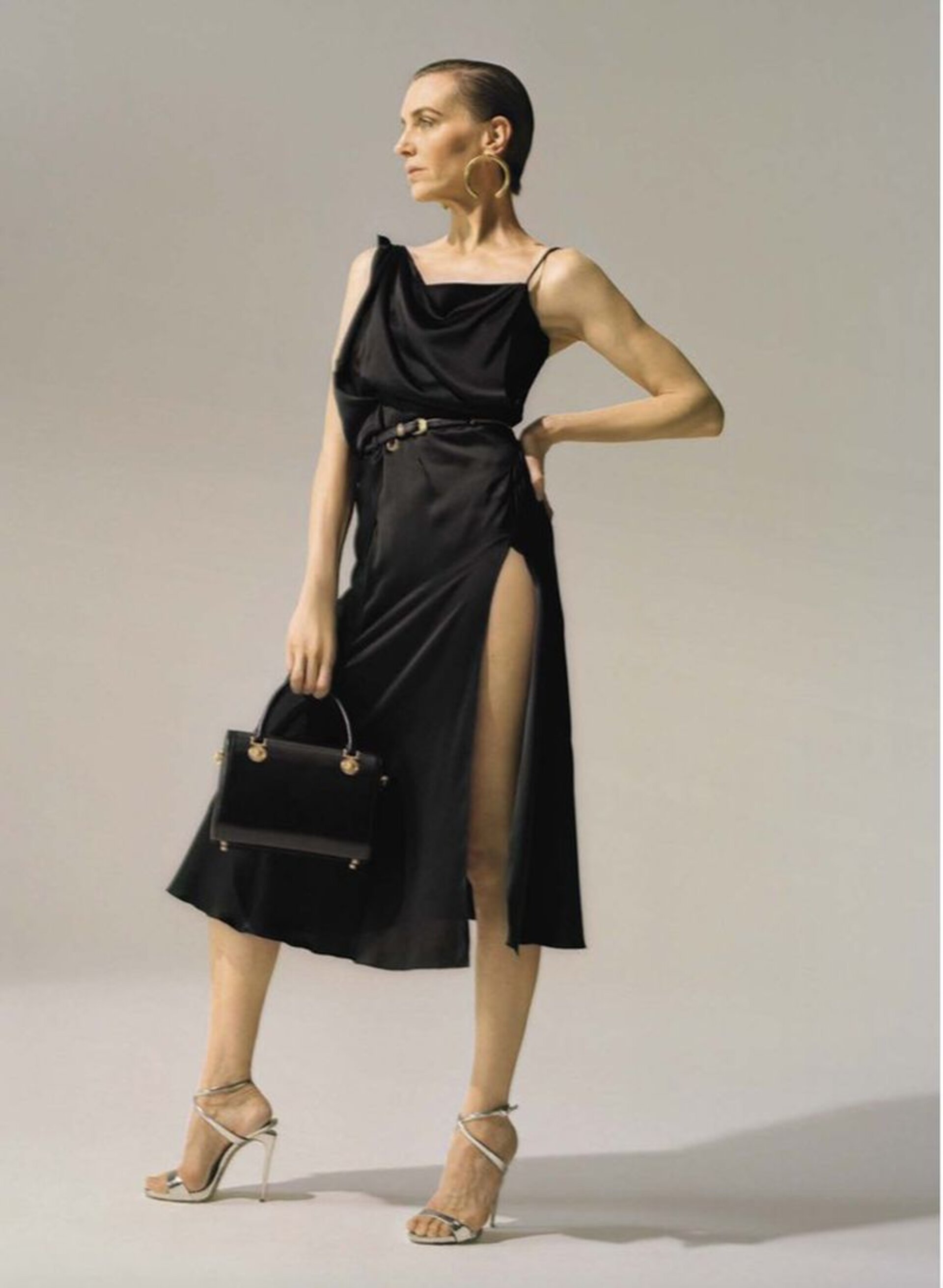 Elegant woman in a black dress and silver heels poses with a black purse against a neutral backdrop, fashion editorial photography.