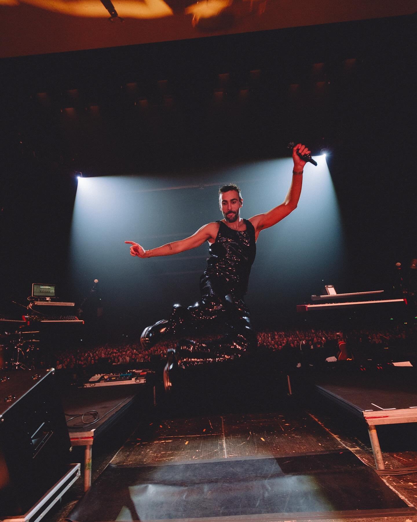 Male musician performing on stage with a microphone, wearing a sparkly black outfit and jumping mid-air during a concert performance.