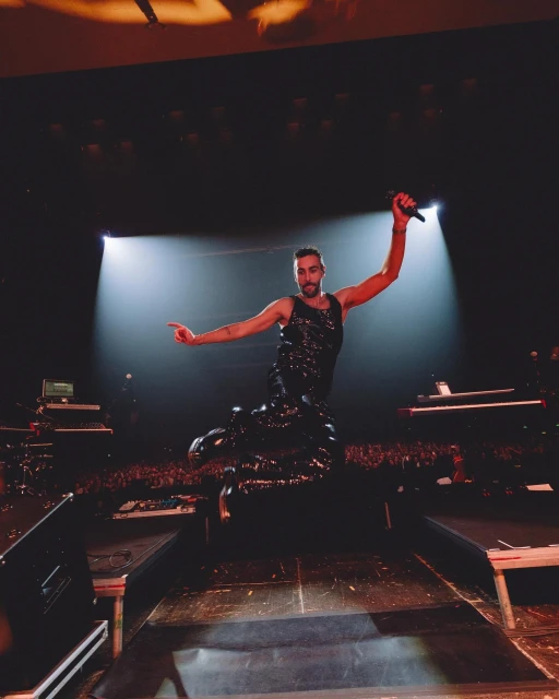 Male musician performing on stage with a microphone, wearing a sparkly black outfit and jumping mid-air during a concert performance.