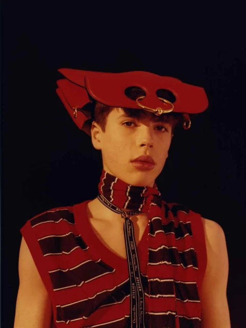 Fashion portrait of a young adult wearing a red striped vest, scarf, and stylized red hat, set against a dark studio background.