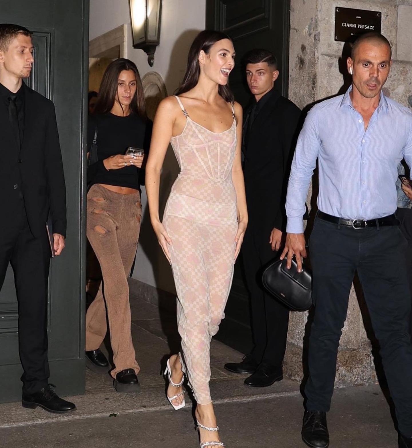 A woman in a sheer dress walks in front of a group of people, with security guards standing around, outside a building in an urban setting.