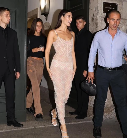 A woman in a sheer dress walks in front of a group of people, with security guards standing around, outside a building in an urban setting.