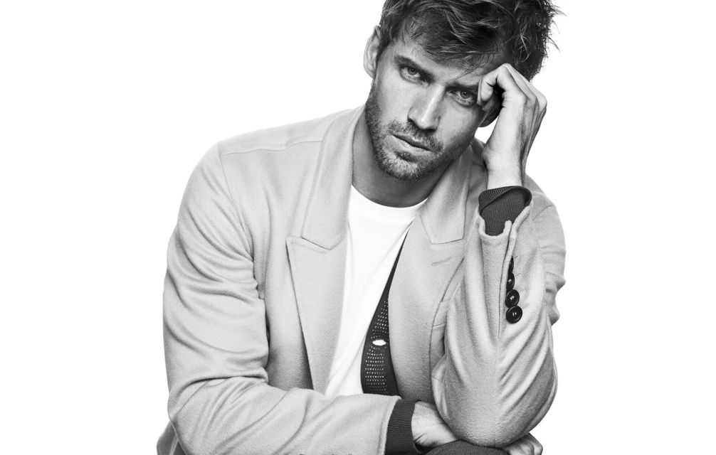 Black-and-white portrait of a handsome adult male model in a blazer and T-shirt, hand to his head, against a white background.
