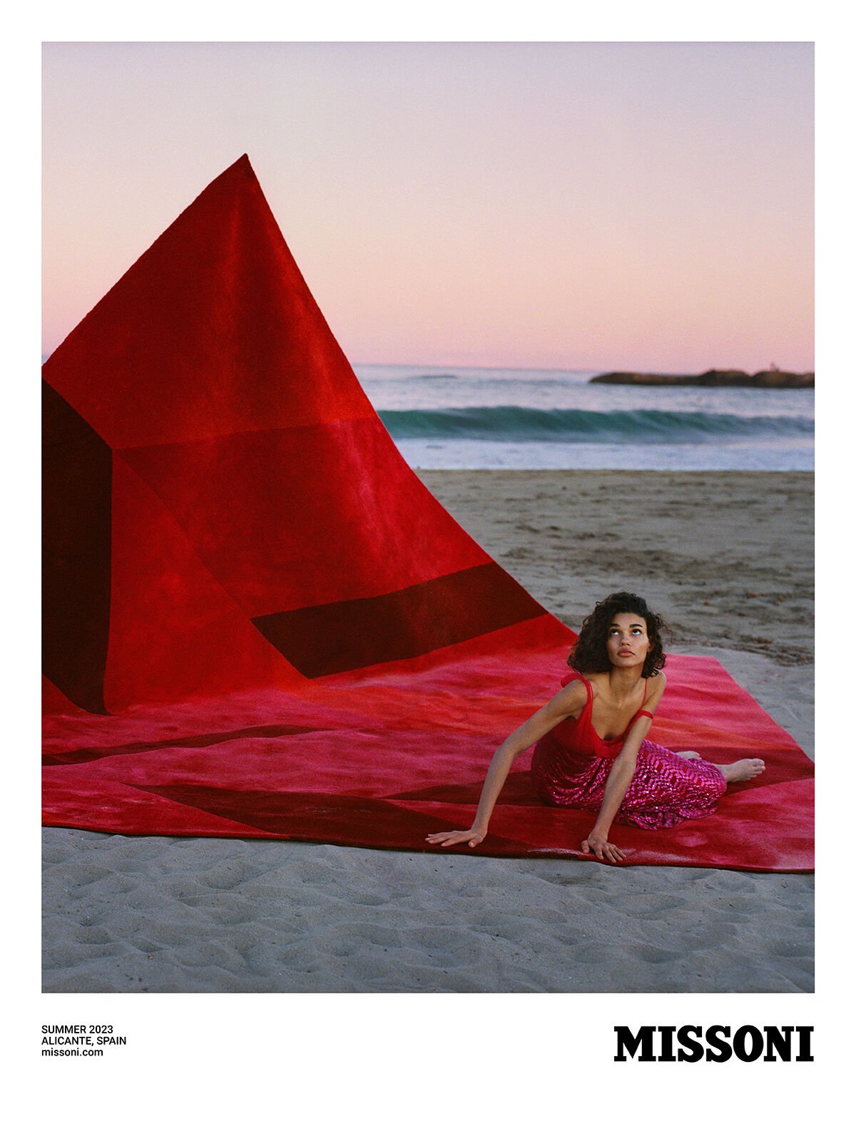 A woman in a red dress poses on a large red rug on a beach with ocean waves and a pink and orange sky at sunset for Missoni.