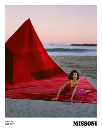 A woman in a red dress poses on a large red rug on a beach with ocean waves and a pink and orange sky at sunset for Missoni.