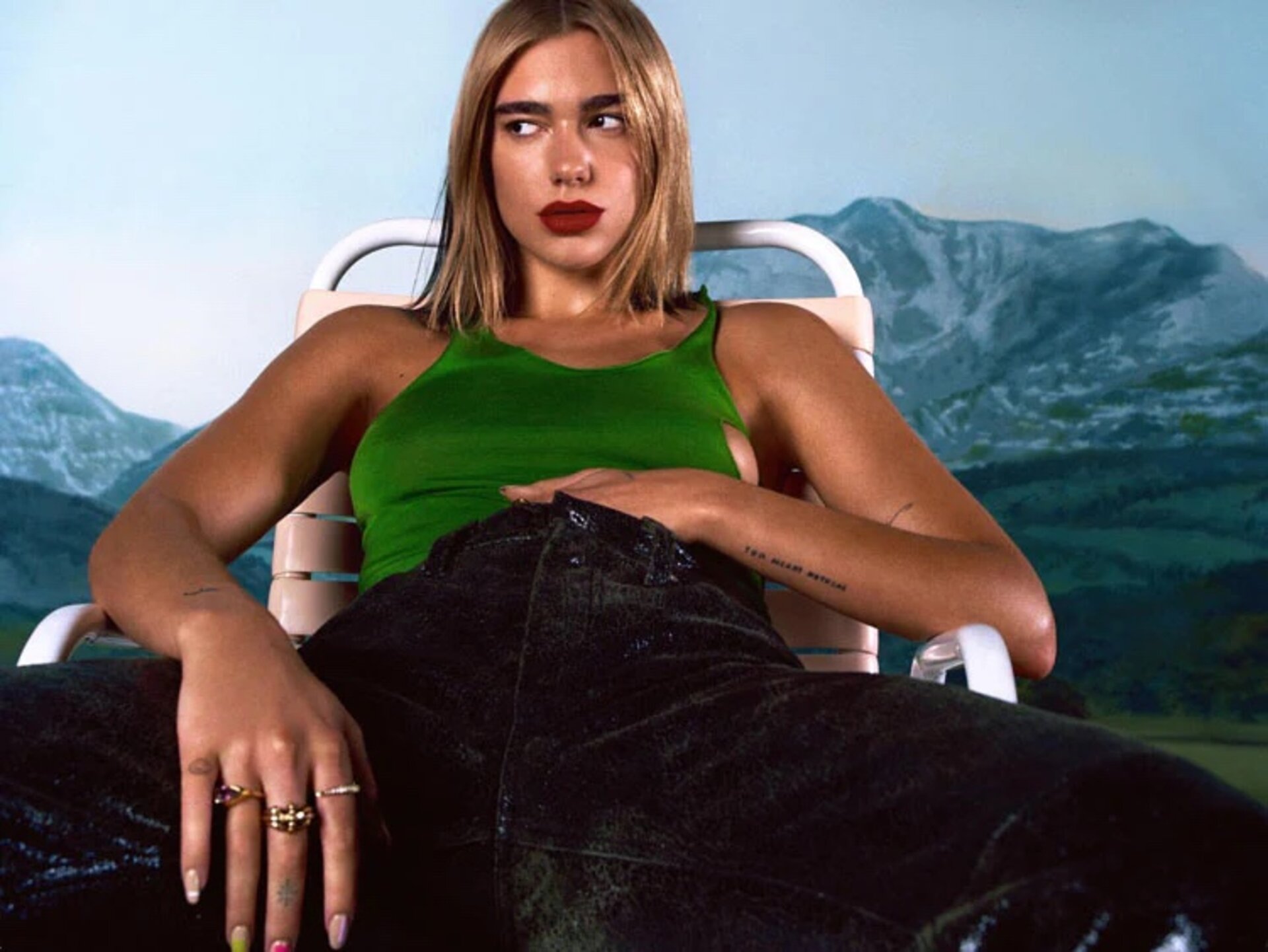 Singer Dua Lipa sits in a chair, wearing a green top and black pants, with a mountain backdrop. She is wearing bright red lipstick.