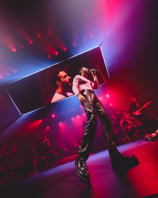 A performer on stage sings into a microphone, wearing leather pants and a pink top, with red and blue stage lighting.