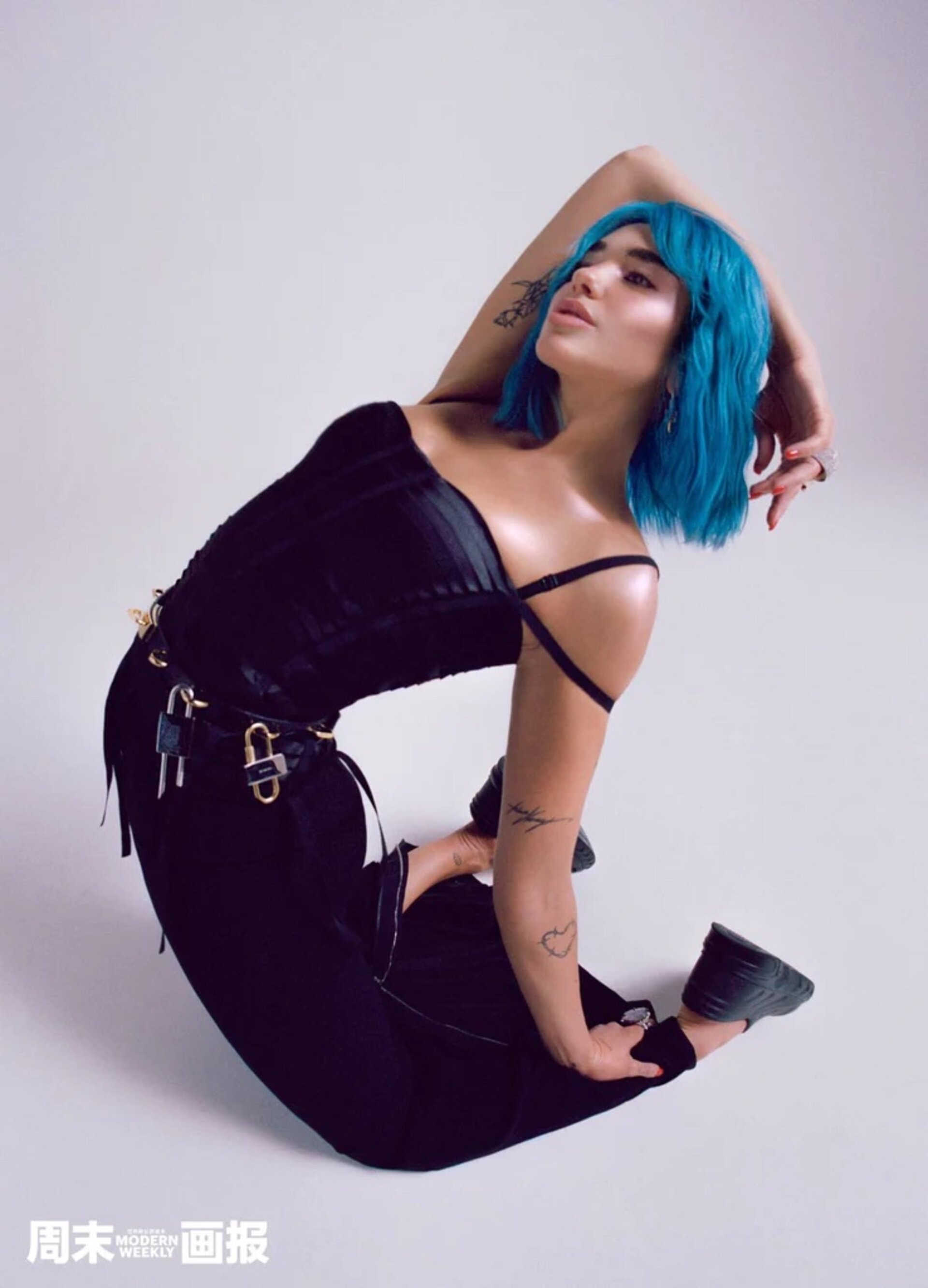 Attractive young woman with blue hair, wearing a black top and pants, poses in a kneeling position against a plain white background.